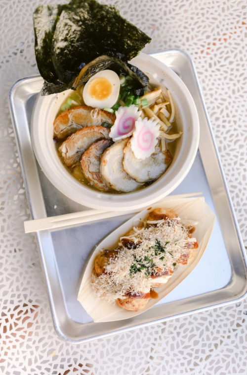 A bowl of ramen with sliced pork, egg, and toppings, alongside a dish of grilled skewers with seasoning on a tray.